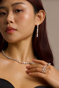 Woman wearing a diamond necklace, drop earrings, and ring against a neutral background